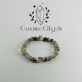 The main product image of the CosmicGlyph Cancer "The Nurturer's Haven" bracelet, featuring a multi-strand design with beads in brown, black, and translucent green tones, laid flat on a light grey background. The CosmicGlyph logo with crystal and leaf motif is displayed above.