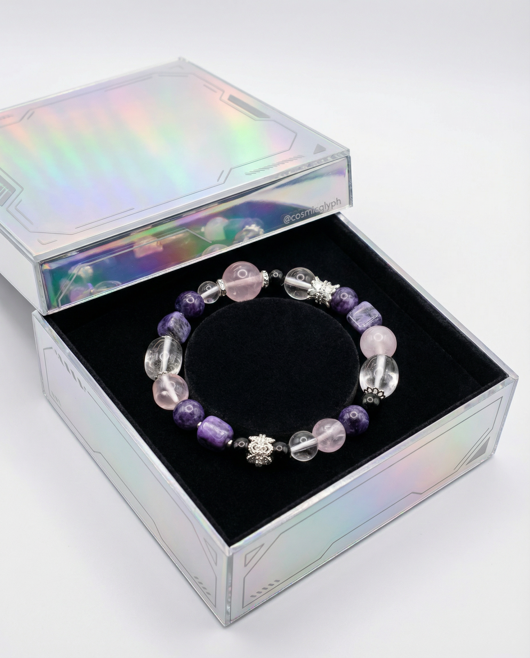 The Aquarius bracelet presented in a square, iridescent transparent box lined with black velvet. The bracelet is composed of purple, pink, clear, and black beads, along with ornate silver beads featuring floral and owl designs. The presentation is clean and artistic. Brand handle: @cosmicglyph.