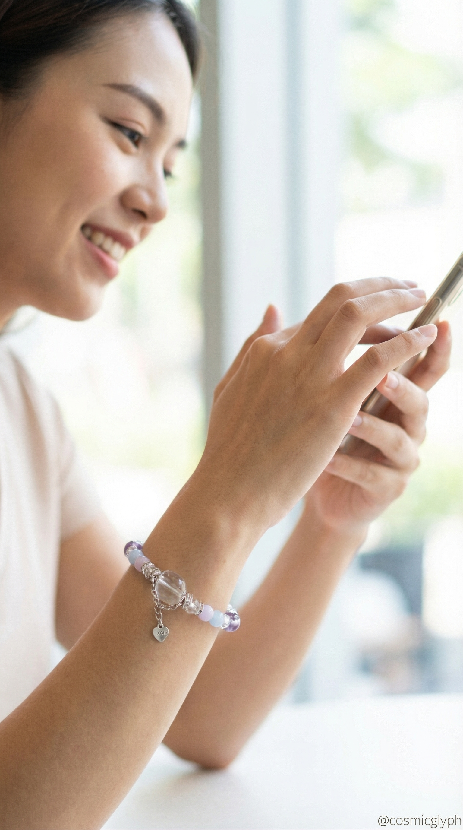 A woman interacts with her phone while wearing the Gemini crystal bracelet. The bracelet's soft purple and pink beads with a heart charm complement the natural, bright setting. The scene captures a moment of modern connection and thoughtful engagement. Text overlay: "LOVE @cosmicglyph".