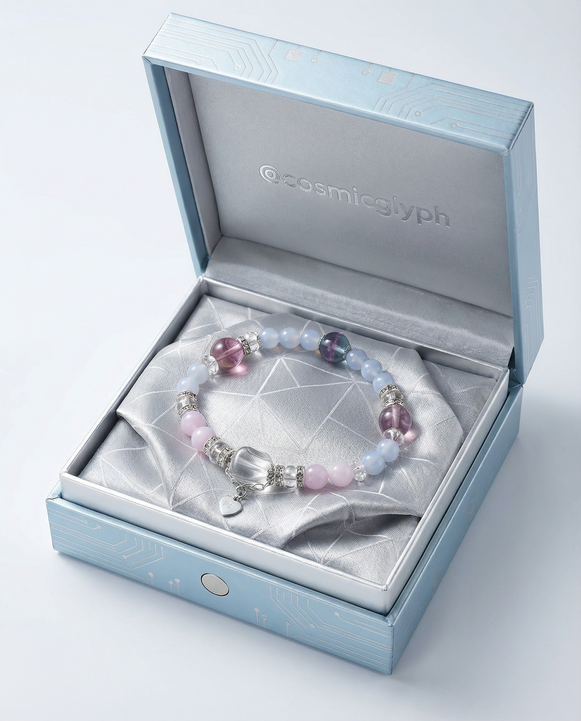 The Gemini bracelet elegantly coiled inside a light blue gift box with a geometric-patterned lining. The bracelet combines soft pink, lavender, and blue beads with a central teardrop crystal. The box is stamped with @cosmicglyph, presenting it as a luxurious gift.