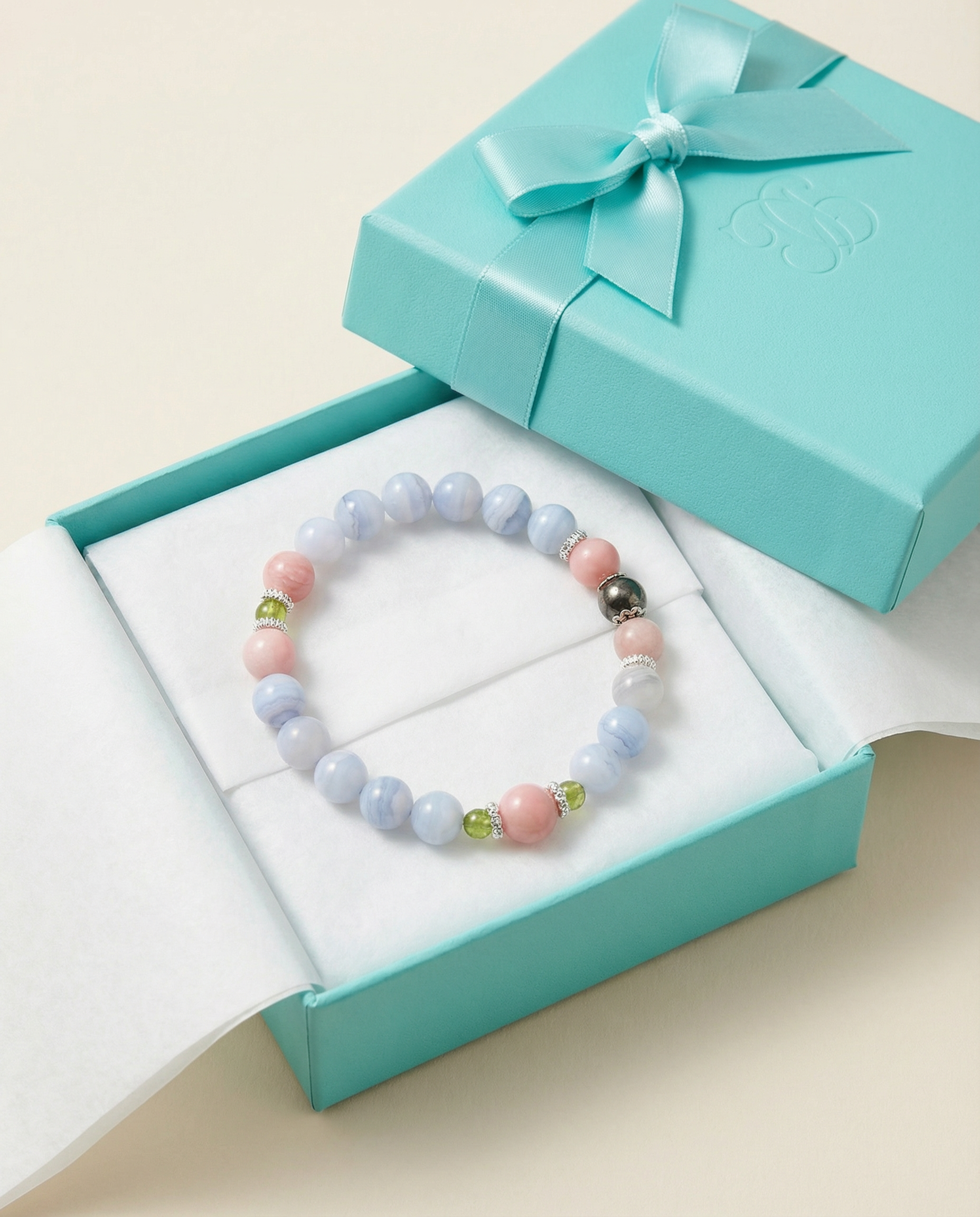 The Libra crystal bracelet nestled inside a light blue gift box with a white flocked lining and a stamped "SS" monogram. The bracelet combines beads of Blue Lace Agate, Pink Opal, Peridot, and Hematite with silver spacers. A同色系 satin bow adorns the lid, presented as a refined gift.